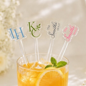 May include: Four clear acrylic drink stirrers in a glass of orange liquid with ice and a mint sprig. The stirrers feature custom text: blue initials LH, green initials KC, black text The Smiths 08.16.27, and pink text The Smiths 08.16.27.