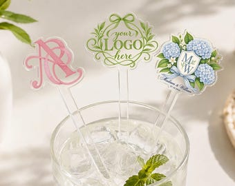 May include: Three clear acrylic drink stirrers in a glass of ice water. One features a pink monogram, one has a green wreath design with the text "your LOGO here", and one displays a blue hydrangea floral crest with "MW" and "Madison & William".