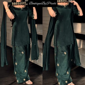 Op de afbeelding: Een donkergroene satijnen salwar kameez-set bestaande uit een tuniek met driekwartmouwen, een bijpassende dupatta en een losvallende broek. De halslijn, mouwboorden en dupatta zijn versierd met gouden borduursels en kleine spiegeltjes. BoutiqueByPreeti.