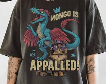 May include: Dark gray t-shirt with a graphic of a blue and red dinosaur and a cat wearing sunglasses and a crown. The text "MONGO IS APPALLED!" is printed on the shirt. The shirt has a Comfort Colors Pepper label.