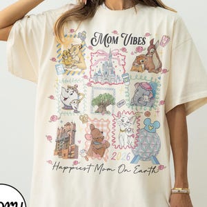 Puede incluir: Camiseta de color marfil con un diseño gráfico colorido que presenta varios personajes de dibujos animados y el texto "Mom Vibes" y "Happiest Mom On Earth". El diseño incluye imágenes de un castillo, animales y el año 2026.