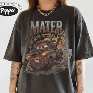 May include: Dark gray t-shirt featuring a graphic of Mater from the movie Cars. The word "MATER" is printed above the image of the character. The shirt also has the words "Comfort Color Pepper" printed on the top left.