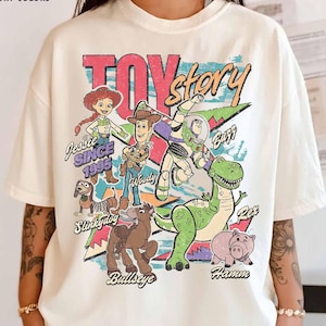 May include: Ivory-colored t-shirt featuring a colorful graphic of Toy Story characters, including Woody, Buzz Lightyear, and Rex. The text "TOY STORY" is prominently displayed, along with "Jessie Since 1995". The shirt has a relaxed fit.