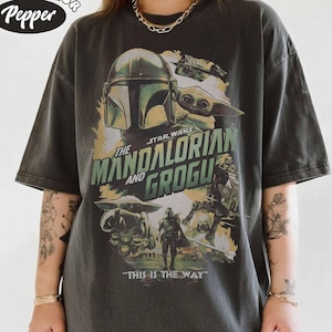 K&ouml;nnte beinhalten: Dunkelgraues T-Shirt mit einem Grafikdesign aus der Star Wars-Serie "The Mandalorian and Grogu". Das Design zeigt Bilder des Mandalorianers, Grogu und anderer Charaktere, mit dem Satz "THIS IS THE WAY" unten. Das Shirt hat das Comfort Colors-Logo.