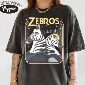 May include: Dark gray t-shirt featuring a cartoon graphic of two zebras with the word "ZEBROS" in yellow. The shirt has a vintage, worn look, with the Comfort Colors and Pepper logos in the upper left corner.