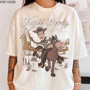 May include: Ivory-colored t-shirt featuring a graphic of Sheriff Woody riding a horse with the text "Sheriff Woody". The design is in shades of brown and tan, with a vintage aesthetic. The shirt is labeled "Comfort Colors".