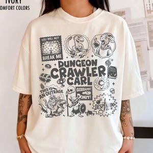 Puede incluir: Una camiseta de manga corta color marfil con un estampado gráfico gris oscuro que incluye el texto Dungeon Crawler Carl. El diseño presenta varias ilustraciones como un dinosaurio, una cabra, un gato sobre una criatura, una calavera y un cofre.
