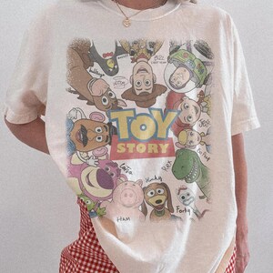 Może przedstawiać: Kremowy t-shirt z kr&oacute;tkim rękawem z dużym, kolorowym nadrukiem graficznym przedstawiającym postacie z Toy Story ułożone w okrąg wok&oacute;ł logo filmu. Wz&oacute;r zawiera postacie takie jak Woody, Buzz Astral, Jessie, Bystry, Rex, Hamm i inne.