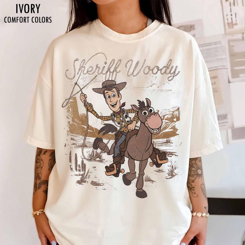 May include: Ivory-colored t-shirt featuring a graphic of Sheriff Woody riding a horse with the text "Sheriff Woody". The design is in shades of brown and tan, with a vintage aesthetic. The shirt is labeled "Comfort Colors".