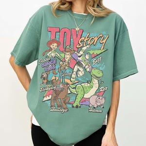 May include: Green t-shirt featuring a graphic of characters from the movie "Toy Story." The design includes Jessie, Woody, Buzz, Rex, Slinkydog, Bullseye, and Hamm, with the text "Toy Story" and "Since 1995."