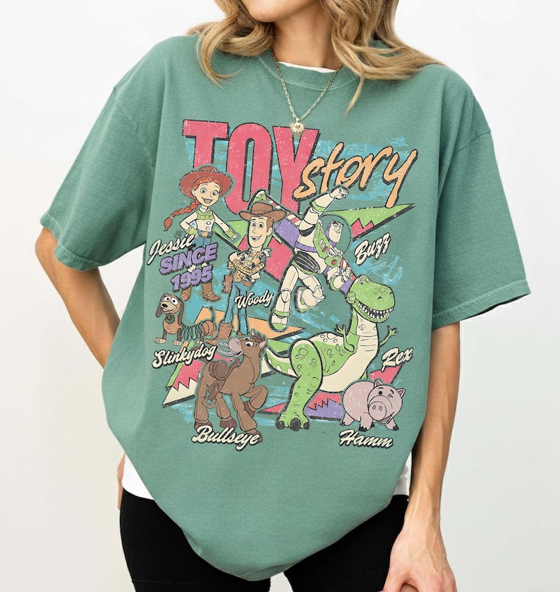 May include: Green t-shirt featuring a graphic of characters from the movie "Toy Story." The design includes Jessie, Woody, Buzz, Rex, Slinkydog, Bullseye, and Hamm, with the text "Toy Story" and "Since 1995."