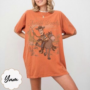 May include: Orange t-shirt featuring a graphic of Sheriff Woody riding a horse, with the text "Sheriff Woody" above the image. The shirt has short sleeves and a relaxed fit.