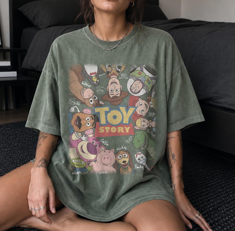 Może przedstawiać: Szałwiowo-zielony t-shirt z kr&oacute;tkim rękawem z dużym nadrukiem graficznym przedstawiającym r&oacute;żne postacie z Toy Story, w tym Chudego, Buzza Astrala, Jessie, Rexa i innych, rozmieszczone wok&oacute;ł centralnego ż&oacute;łto-czerwonego logo Toy Story.