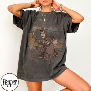 May include: Dark grey t-shirt featuring a graphic of Sheriff Woody riding a horse, with the text "Sheriff Woody" above the image. The design has a vintage, distressed look.