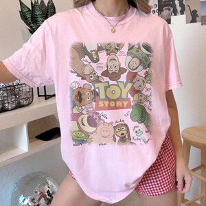 Może przedstawiać: Jasnor&oacute;żowy t-shirt z kr&oacute;tkim rękawem z dużą grafiką na środku przedstawiającą r&oacute;żne postacie z Toy Story ułożone w okrąg wok&oacute;ł logo Toy Story. Wśr&oacute;d postaci znajdują się Woody, Buzz Astral, Jessie, Rex, Hamm, Cienki i inne.