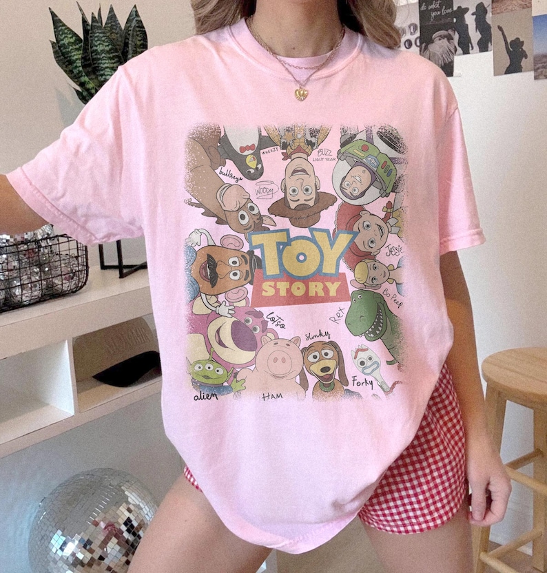 Może przedstawiać: Jasnor&oacute;żowy t-shirt z kr&oacute;tkim rękawem z dużą grafiką na środku przedstawiającą r&oacute;żne postacie z Toy Story ułożone w okrąg wok&oacute;ł logo Toy Story. Wśr&oacute;d postaci znajdują się Woody, Buzz Astral, Jessie, Rex, Hamm, Cienki i inne.