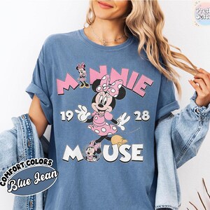 May include: Blue jean Comfort Colors t-shirt featuring a Minnie Mouse graphic. The design includes the word "MINNIE" in pink block letters, the year "1928", and the word "MOUSE". Minnie Mouse is depicted in a pink polka dot dress.