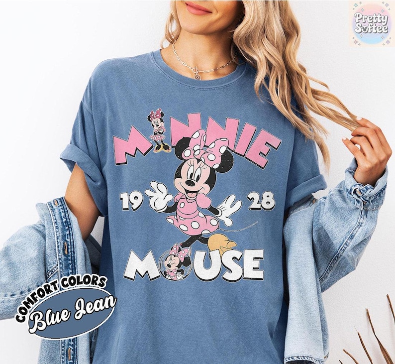 May include: Blue jean Comfort Colors t-shirt featuring a Minnie Mouse graphic. The design includes the word "MINNIE" in pink block letters, the year "1928", and the word "MOUSE". Minnie Mouse is depicted in a pink polka dot dress.