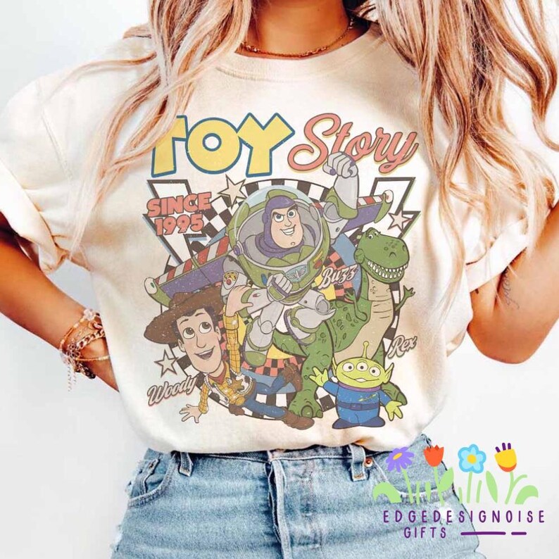 K&ouml;nnte beinhalten: Cremefarbenes T-Shirt mit einem Grafikdesign im Vintage-Stil von Toy Story-Charakteren, darunter Woody, Buzz Lightyear, Rex und die kleinen gr&uuml;nen M&auml;nnchen. Das Shirt tr&auml;gt den Text "Toy Story Since 1995" in einer Retro-Schriftart.
