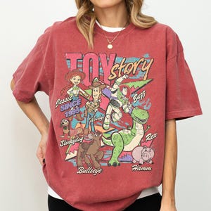 May include: Red t-shirt featuring a graphic of Toy Story characters including Woody, Buzz Lightyear, and Jessie. The design includes the text "Toy Story" and "Since 1995". The shirt has a crew neck and short sleeves.