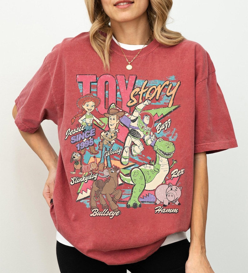 May include: Red t-shirt featuring a graphic of Toy Story characters including Woody, Buzz Lightyear, and Jessie. The design includes the text "Toy Story" and "Since 1995". The shirt has a crew neck and short sleeves.