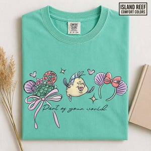 May include: A seafoam green t-shirt featuring a colorful graphic of Flounder, a seashell, and a headband with a bow. The text "Part of your world" is below the graphic. The shirt has a crew neck and the brand name "Island Reef" is in the upper right corner.