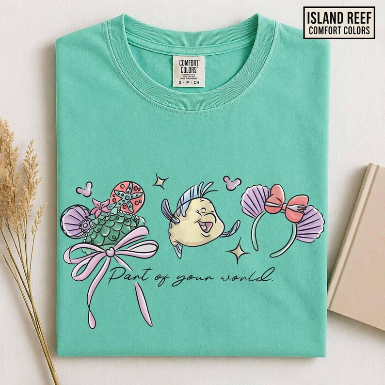 May include: A seafoam green t-shirt featuring a colorful graphic of Flounder, a seashell, and a headband with a bow. The text "Part of your world" is below the graphic. The shirt has a crew neck and the brand name "Island Reef" is in the upper right corner.