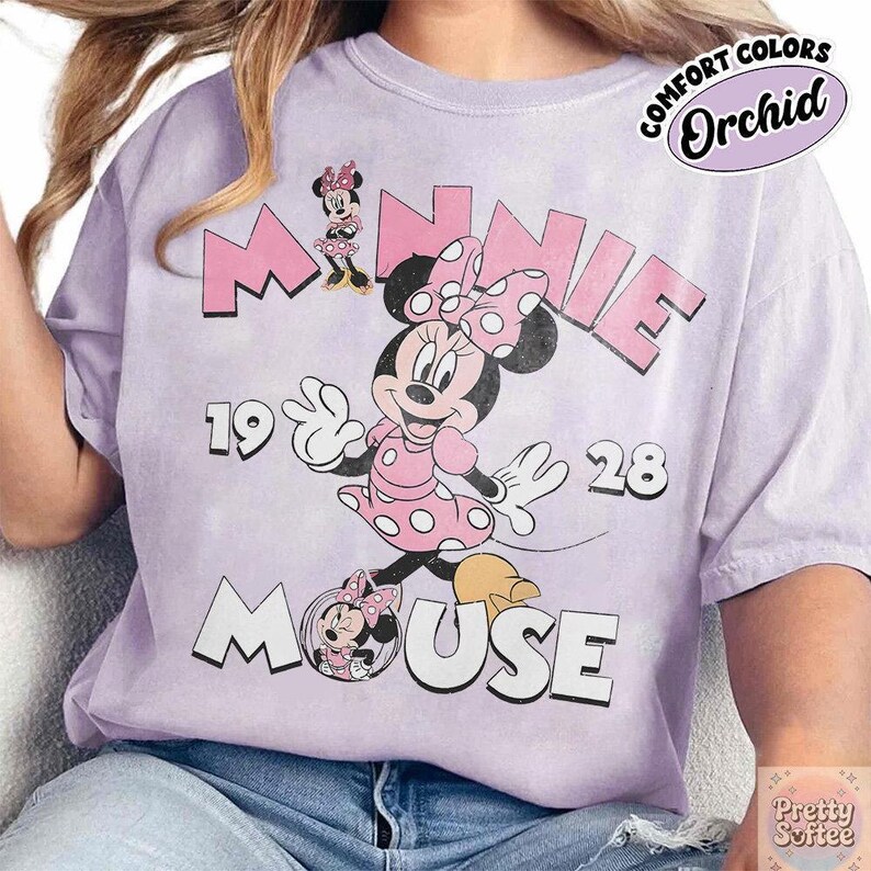 May include: Orchid-colored t-shirt featuring Minnie Mouse. The design includes the word "MINNIE" in pink, the year "1928", and the word "MOUSE". Minnie Mouse is depicted in a pink polka-dot dress and bow.