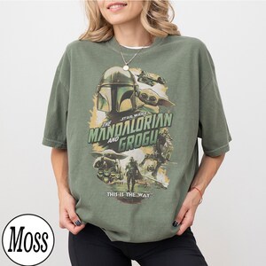 K&ouml;nnte beinhalten: Olivgr&uuml;nes T-Shirt mit einem Grafikdesign von The Mandalorian und Grogu. Das Design zeigt den Helm des Mandalorianers, Grogu und den Text "This is the Way". Das T-Shirt hat kurze &Auml;rmel und eine lockere Passform.