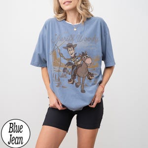 May include: A blue t-shirt featuring a graphic of Sheriff Woody riding a horse with the text "Sheriff Woody". The shirt has a vintage, distressed look. The shirt is a light blue color.