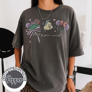 May include: Dark grey t-shirt featuring a colorful graphic of cartoon characters and the text "Part of your world." The design includes a bow, a fish, and mouse ears. The shirt is from Comfort Colors.