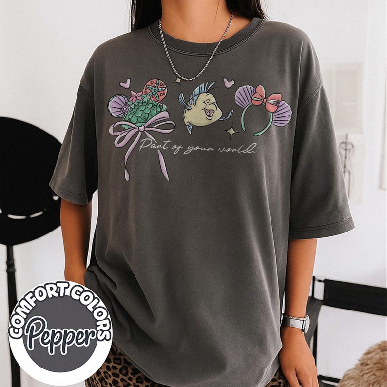 May include: Dark grey t-shirt featuring a colorful graphic of cartoon characters and the text "Part of your world." The design includes a bow, a fish, and mouse ears. The shirt is from Comfort Colors.