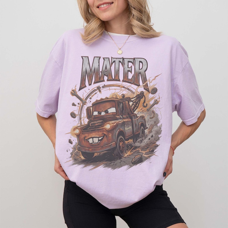 May include: A light purple short-sleeve t-shirt featuring a graphic print of a brown tow truck character with the text MATER above it. The design includes illustrations of mechanical tools, dust clouds, and debris surrounding the vehicle.
