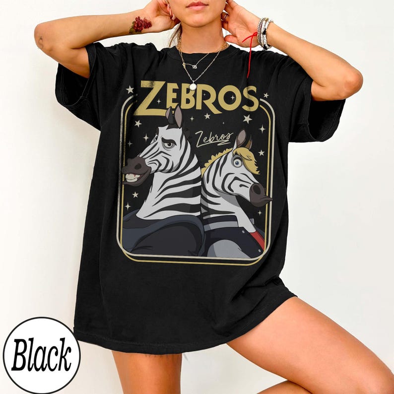 May include: Black t-shirt featuring a cartoon illustration of two zebras with the text "ZEBROS" in gold lettering. The zebras are in a rectangular frame with stars. The word "Black" is in a white circle at the bottom.