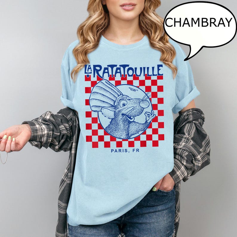 May include: Light blue chambray t-shirt featuring a graphic of Remy from the movie Ratatouille. The design includes the movie title, a red and white checkered pattern, and the text "Paris, FR".