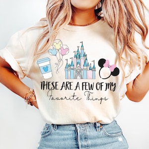 May include: Cream-colored t-shirt featuring a graphic design with a coffee cup, balloons, a castle, and a Minnie Mouse silhouette. The text "These are a few of my Favorite Things" is printed below the graphics.