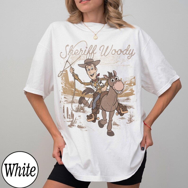 May include: White t-shirt featuring a graphic of Sheriff Woody riding a horse, with the text "Sheriff Woody" in a vintage style. The design has a sepia-toned background with desert elements. The shirt is a casual, oversized fit.