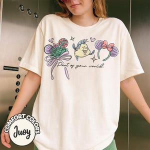 May include: Cream-colored t-shirt featuring a colorful graphic of sea-themed characters and the text "Part of your world." The design includes a mermaid tail, a yellow fish, and a headband with a bow. The shirt has short sleeves and a round neckline.