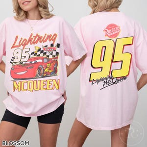 May include: Light pink t-shirt featuring Lightning McQueen from the Cars movie. The front graphic shows McQueen with the number 95, a checkered flag, and the text "Lightning McQueen". The back displays a large yellow "95" and the text "Lightning McQueen".