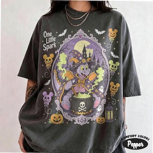 Puede incluir: Una camiseta gris carbón con un gráfico de un dragón morado con sombrero de bruja revolviendo un caldero. El diseño incluye el texto One Little Spark, calabazas, murciélagos y globos con forma de ratón dentro de un marco morado ornamentado.