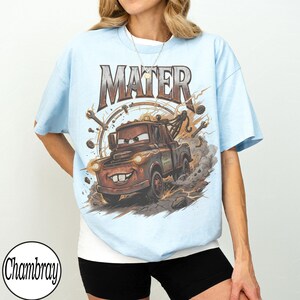 May include: Light blue t-shirt featuring a graphic of Mater from the movie Cars. The word "MATER" is above the character. The graphic shows Mater with a big smile, surrounded by tools and lightning bolts. The shirt is a light blue color.