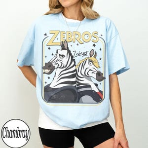 May include: Light blue t-shirt featuring a cartoon graphic of two zebras with the text "Zebros". The zebras are black and white with cartoonish expressions. The shirt has a crew neck and short sleeves.