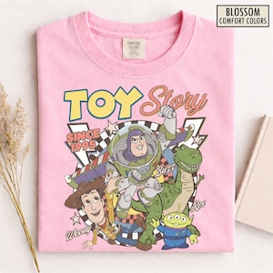 K&ouml;nnte beinhalten: Hellrosa T-Shirt mit einer Grafik von Toy Story-Charakteren, darunter Buzz Lightyear, Woody und Rex. Das Design enth&auml;lt den Text "TOY Story" und "SINCE 1995". Das T-Shirt besteht aus Comfort Colors-Material.