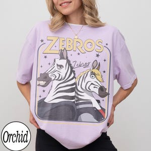 May include: A light purple t-shirt featuring a cartoon graphic of two zebras with the text "ZEBROS" in yellow. The zebras are wearing jackets and have black and white stripes. The shirt has a relaxed fit.