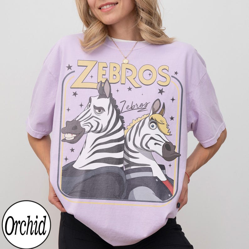 May include: A light purple t-shirt featuring a cartoon graphic of two zebras with the text "ZEBROS" in yellow. The zebras are wearing jackets and have black and white stripes. The shirt has a relaxed fit.