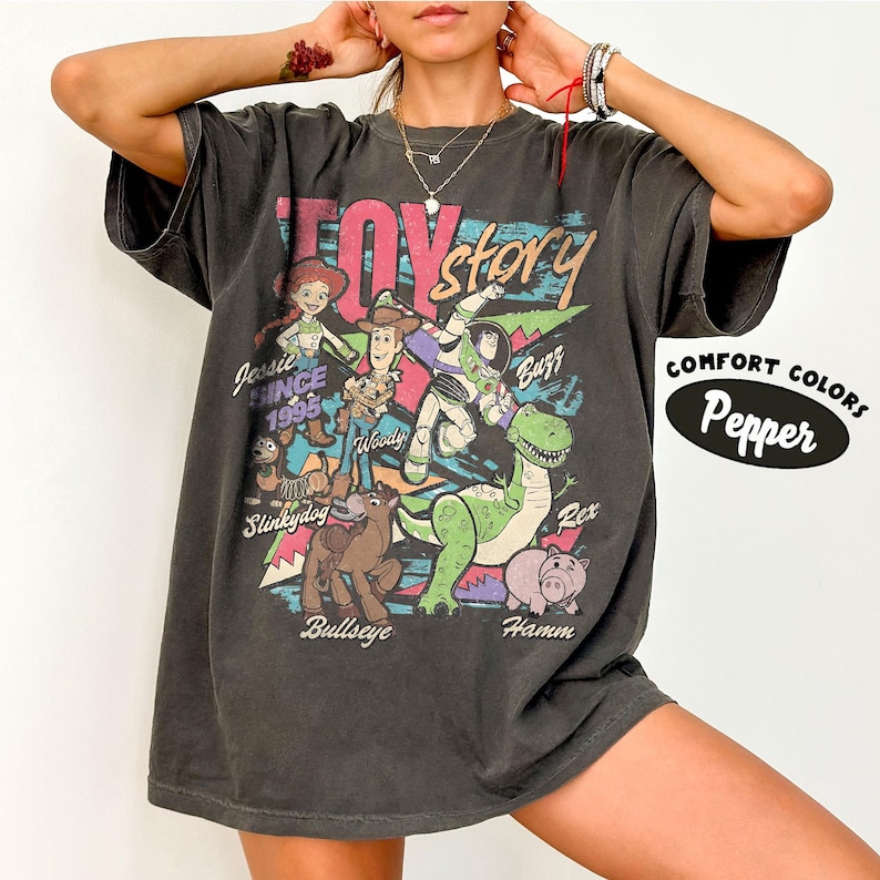 May include: Dark gray t-shirt featuring a colorful graphic of Toy Story characters, including Woody, Buzz Lightyear, and Jessie. The text "Toy Story" is in a retro font, with "Since 1995" below Jessie. The shirt is labeled "Comfort Colors Pepper".