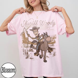 May include: A light pink t-shirt featuring a graphic of Sheriff Woody riding a horse, with the text "Sheriff Woody" in a glittery font. The design is set against a desert landscape.