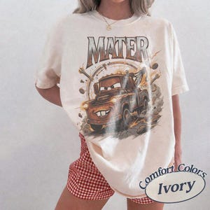 May include: An ivory short-sleeve t-shirt featuring a graphic print of the character Mater from the movie Cars. The design includes the text MATER in a metallic font above the brown tow truck, surrounded by mechanical tools and dust clouds.