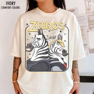May include: Ivory-colored t-shirt with the word "ZEBROS" in yellow, featuring cartoon zebras. The shirt has the text "IVORY COMFORT COLORS" at the top. The zebras are in a rectangular frame with stars.