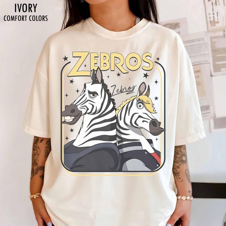 May include: Ivory-colored t-shirt with the word "ZEBROS" in yellow, featuring cartoon zebras. The shirt has the text "IVORY COMFORT COLORS" at the top. The zebras are in a rectangular frame with stars.